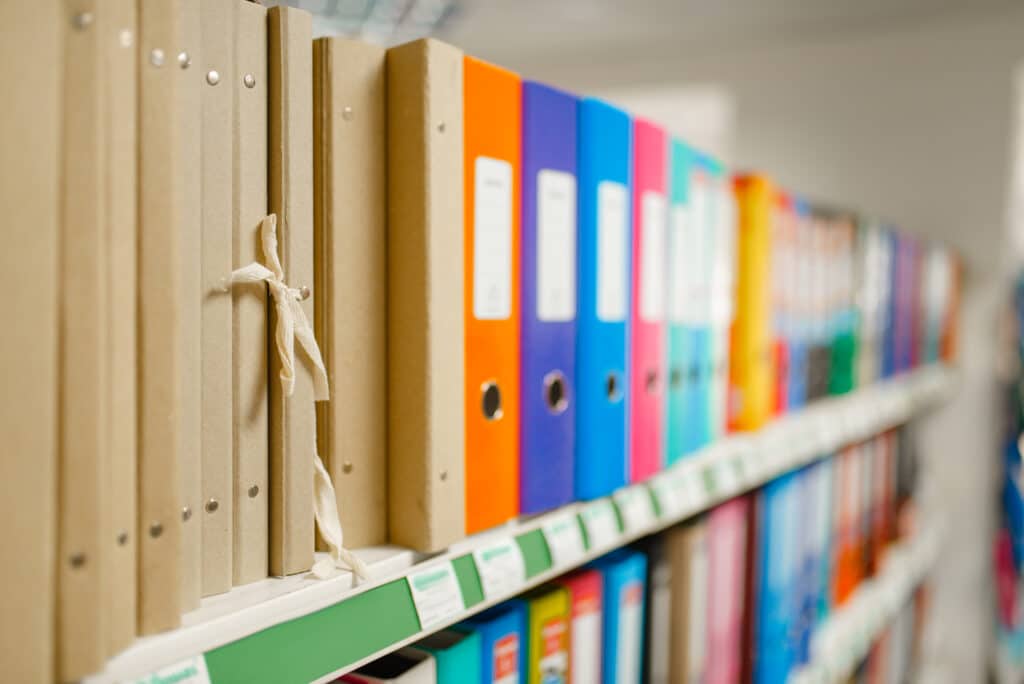 Ein Regal mit verschiedenen Ordnern in bunten Farben, darunter braune und farbige Aktenordner, die ordentlich aufgereiht sind. Im Vordergrund ist ein brauner Ordner mit einer Schnur sichtbar. Ideal für Büroorganisation und Dokumentenmanagement.