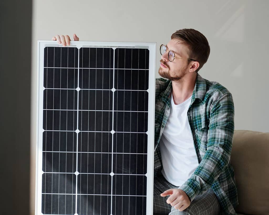 Mann mit Brille sitzt auf einem Sofa und hält ein Solarpanel in der Hand, umgeben von einem hellen Raum.
